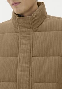 Tarnfarbene Cord-Pufferjacke mit hohem Kragen, gestepptem Material und vertikaler Rippung, mit Nahtdetails an der Vorderseite.