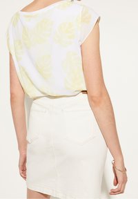 Camisa blanca con patrón de hojas amarillas; mangas cortas; combinada con una falda de denim blanca con diseño de cinco bolsillos. Pulsera dorada a la vista.