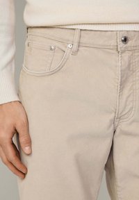 Pantalon en velours côtelé beige clair avec rivets en métal et détails de couture, porté avec un pull côtelé crème, montrant une main détendue près de la hanche.