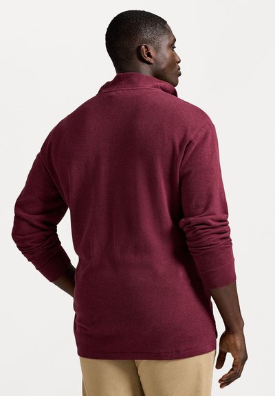 Man facing sideways wears long-sleeve burgundy sweater and beige pants with one hand on hip against plain white background.