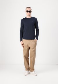 Camicia a maniche lunghe blu marina con un piccolo logo, abbinata a pantaloni beige e sneakers bianche. Il modello indossa anche occhiali da sole neri.