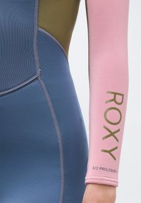 Κοντινή λήψη στο πλάι και το χέρι μιας στολής κατάδυσης, που δείχνει μπλε, ελιάς και ροζ πάνελ με τα κείμενα "ROXY" και "3/2 PROLOGUE+" στο ροζ μανίκι.