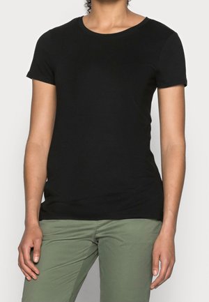 Person trägt ein eng anliegendes schwarzes T-Shirt mit kurzen Ärmeln und olivgrüne Hosen, steht vor einem einfarbigen hellen Hintergrund.