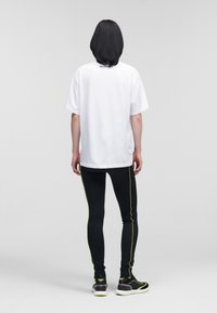 Witte oversized t-shirt met korte mouwen en merklogo op de achterkant, gecombineerd met zwarte leggings met groene zijstrepen en zwarte sportschoenen.