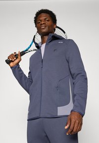 Homme en survêtement bleu tenant une raquette de tennis sur l'épaule, debout devant un fond clair uni.