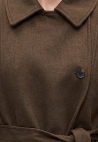 Manteau en laine marron avec un large col, un bouton noir visible unique et une ceinture nouée à la taille sur une personne.