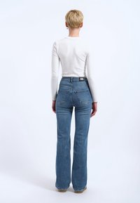 Personne aux cheveux blonds courts portant un haut blanc à manches longues et un jean bleu évasé taille haute, debout de dos sur un fond blanc.