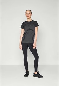 Kvinna som står i en mörk atletisk t-shirt, svarta leggings och svarta löparskor framför en enkel ljus bakgrund.