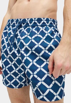 Torso maschile che indossa pantaloncini da nuoto blu con motivo a reticolo geometrico bianco e vita regolabile con coulisse.