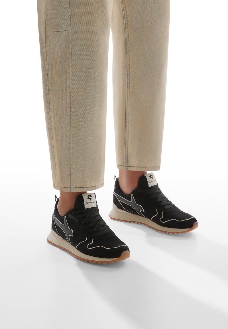 Personne portant un pantalon beige court et des baskets noires avec des semelles blanches et marron clair, debout sur une surface blanche.