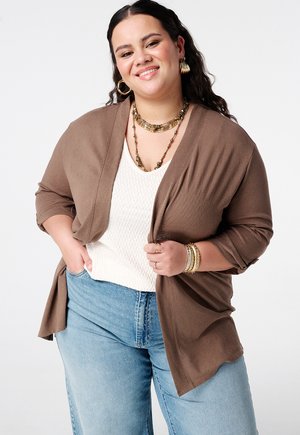 Femme souriante aux cheveux bouclés foncés portant des bijoux en or, un cardigan marron, un haut blanc texturé et un jean bleu clair sur un fond uni.