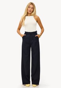 Débardeur blanc côtelé associé à un pantalon large taille haute bleu marine. Le mannequin se tient les mains dans les poches, portant des escarpins jaunes à bouts pointus.