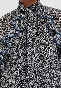 Svart och vit blommönstrad blus med volanger och blå detaljer vid halsringningen, tillverkad av ett genomskinligt tyg. Har en rynkad krage.