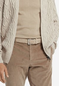 Beige stickad kofta med ett kabelmönster, lagd över en ljusbeige skjorta, i kombination med kordflöjtbyxor och ett flätat bälte.