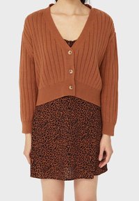 Verkort cardigan in roestkleur met geribbelde structuur, V-hals en vier houten knopen over een zwarte jurk met luipaardprint.