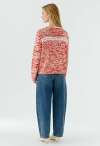 Frau mit blondem Haar, die einen rot-weiß gemusterten Pullover, blaue weit geschnittene Jeans und weiße Schuhabsätze trägt, mit dem Rücken zum Betrachter vor weißem Hintergrund.