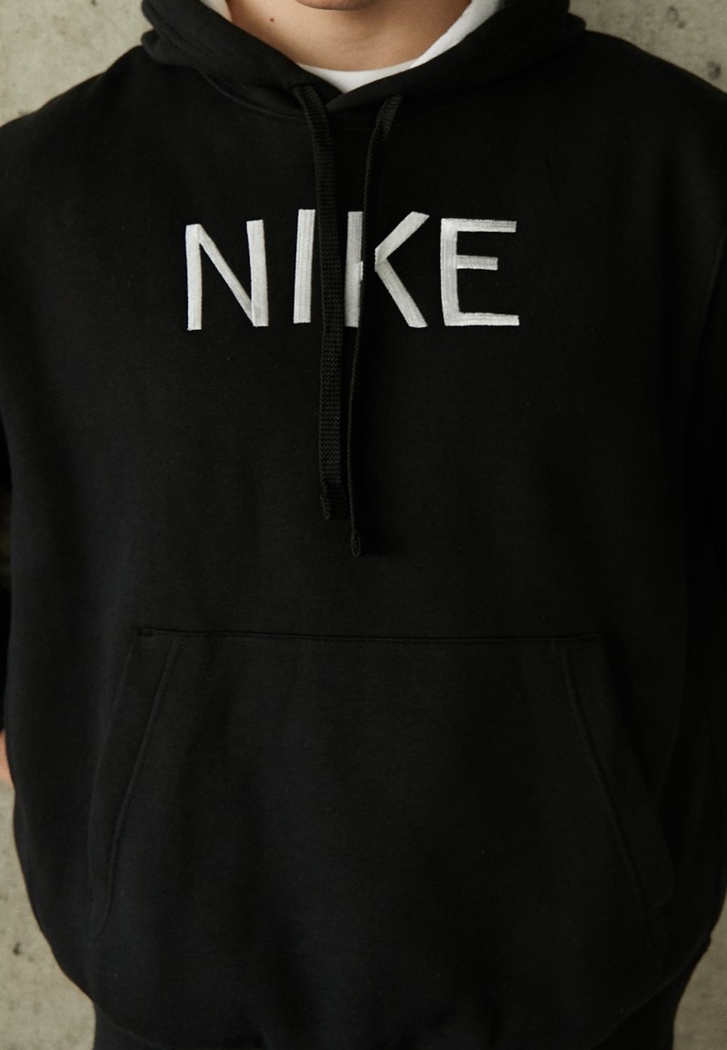 Sweat à capuche noir avec logo "NIKE" blanc, capuche avec cordon, poche avant, texture douce, poignets et ourlet côtelés. Design simple et décontracté.