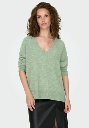 Pull en maille léger, vert clair, avec un décolleté en V profond et des manches courtes roulées, associé à une jupe noire avec une fente sur le côté.