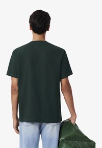 T-shirt in cotone verde con maniche corte, scollo rotondo e vestibilità rilassata. Il modello tiene in mano una giacca a vento verde. Jeans leggeri blu visibili.