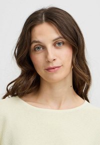 Hellgelber, strukturierter Pullover mit weitem Rundhalsauschnitt. Das Modell hat mittelbraunes, gewelltes Haar und klare blaue Augen. Sauberer Hintergrund.