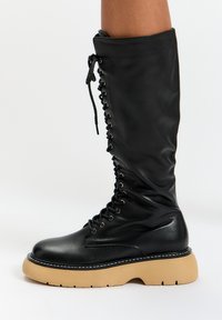 Stivale nero al ginocchio con finitura lucida e texture, caratterizzato da lacci frontali e una robusta suola platform beige con cuciture bianche.