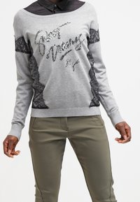 Sweatshirt en tricot gris avec des panneaux latéraux en dentelle noire et un design de texte en sequins. Assorti à un pantalon vert olive, mettant en avant une coupe décontractée.