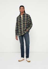 Camisa de cuadros verde y relajada, combinada con jeans azul oscuro y zapatillas beige sobre un fondo liso.
