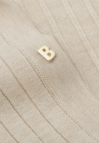 Beżowa dzianina z pionowym prążkowanym wzorem, z małym złotym metalowym akcentem w kształcie litery "B" osadzonym z boku.