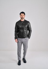 Černá kožená bomber bunda s hladkou texturou, zapínáním na zip vpředu a úzkým střihem, kombinovaná se světle šedými kalhotami a černými kotníkovými botami.