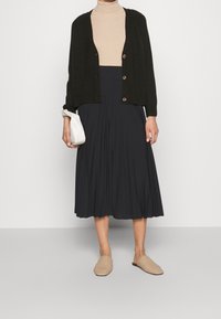Czarny kardigan z zapięciem na guziki, beżowy golf, ciemna plisowana spódnica oraz plecione beżowe mule. Biała kopertówka w ręku.