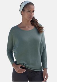 Langärmliges grünes Oberteil aus weichem Stoff mit weitem Ausschnitt und lässiger Passform, kombiniert mit dunklen Jeans und einem hellblauen Haarband.