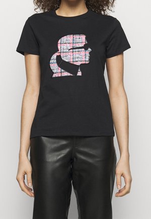 Kvinde iført en sort t-shirt med et pink og hvidt ternet silhuetgrafik af en persons profil foran, parret med sorte læderbukser.