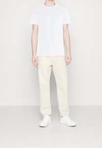Uomo vestito con una T-shirt bianca a maniche corte, pantaloni dritti bianco sporco e sneakers bianche in piedi contro uno sfondo chiaro e uniforme.