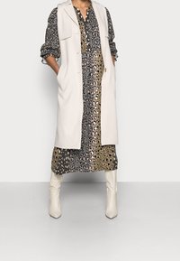 Beżowa, bezrękawnikowa kamizelka nałożona na sukienkę w wielokolorowy wzór panterki; długość do kolan, z luźnymi rękawami, zestawiona z białymi botkami na obcasie.