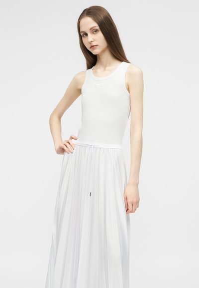 Jeune femme aux longs cheveux bruns portant une robe blanche sans manches avec un corsage ajusté et une jupe plissée, debout devant un fond uni.