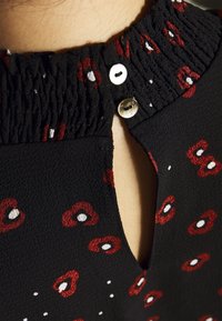 Blusa nera con scollo smock, una apertura a forma di chiave sulla schiena e un motivo di forme astratte rosse e bianche. Include un dettaglio con bottone.