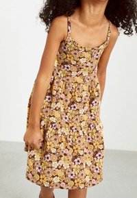 Niña con un vestido sin mangas marrón con patrones florales en amarillo, blanco y rosa, de pie frente a un fondo claro y liso.