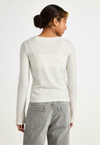 Langærmet ribbet top i lys beige med rund hals. Har fitted ærmer og en glat tekstur, båret med grå afslappede jeans.