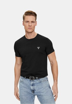 Uomo con t-shirt nera a maniche corte con piccolo logo sul petto e jeans azzurri chiari, in piedi contro uno sfondo grigio chiaro uniforme.