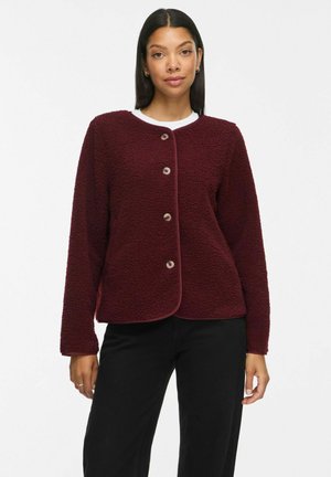 Femme portant une veste polaire bordeaux boutonnée à manches longues et un pantalon noir, debout devant un fond blanc.