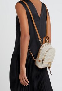 Beige reppuluku, jossa on monogrammikuosi, kultaiset yksityiskohdat ja ruskeat aksentit. Ominaisuuksiin kuuluu kaksi vetoketjullista lokeroa ja säädettävät hihnat.