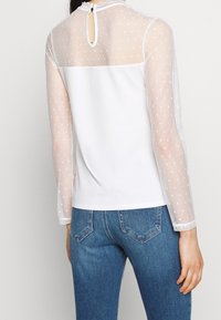 Top bianco con maniche lunghe trasparenti a pois e retro solido, con un'apertura a chiave sul collo. Indossato con jeans in denim blu.