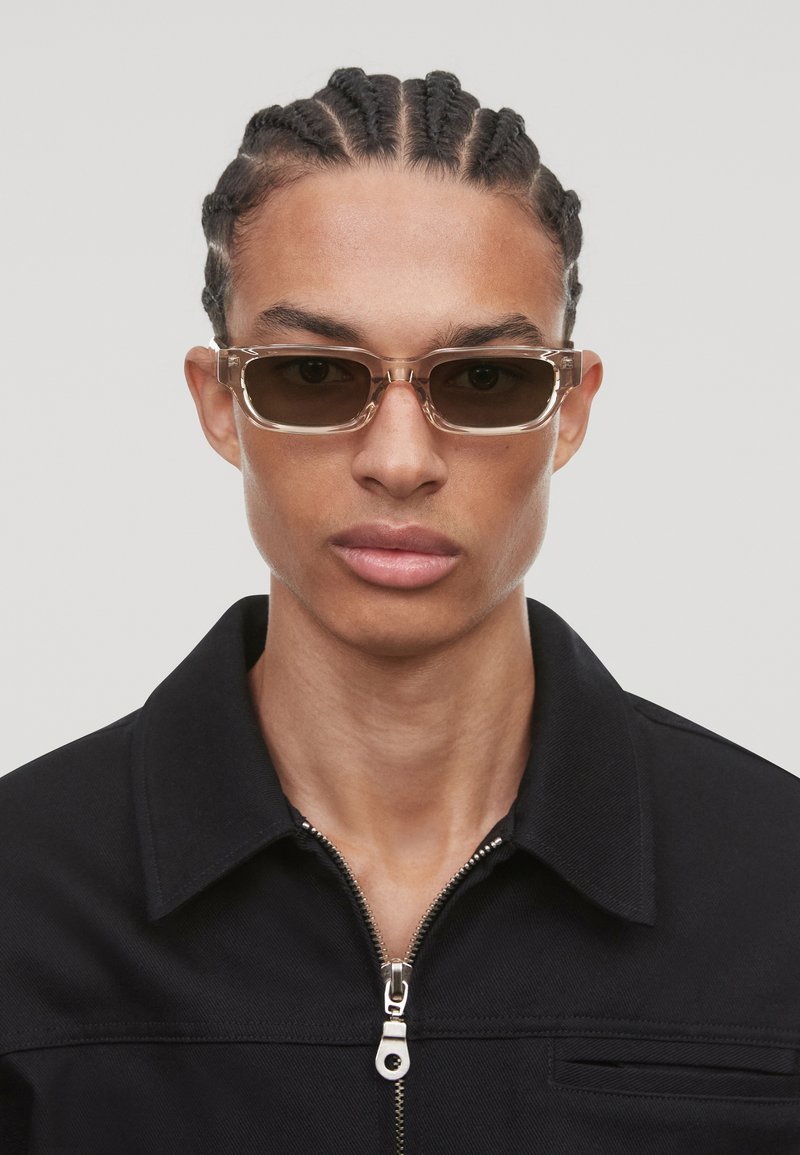 Lunettes de soleil avec une monture transparente et des verres foncés, de forme rectangulaire, dotées de branches fines, portées par une personne dans une veste noire zippée.