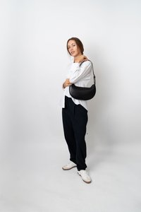 Kobieta w białej koszuli, czarnych spodni w prążki i sneakersach trzymająca czarną torebkę na ramię na jednolitym białym tle.