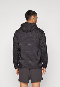 Svart hettegenser med teksturert mønster, lett stoff, lange ermer og reflekterende detaljer. Båret over grå shorts.