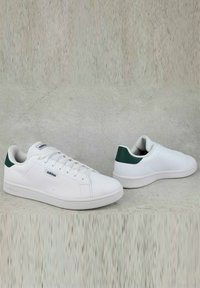 Scarpe da ginnastica bianche con una finitura in pelle liscia, accenti verdi sul tallone, design con lacci piatti e suola in gomma testurizzata. Logo del marchio ben visibile sul lato.