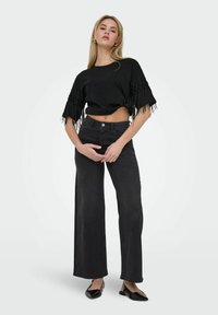 Un haut noir court avec des manches à franges associé à un jean noir taille haute à jambes larges. Le modèle porte des ballerines noires. Fond simple.