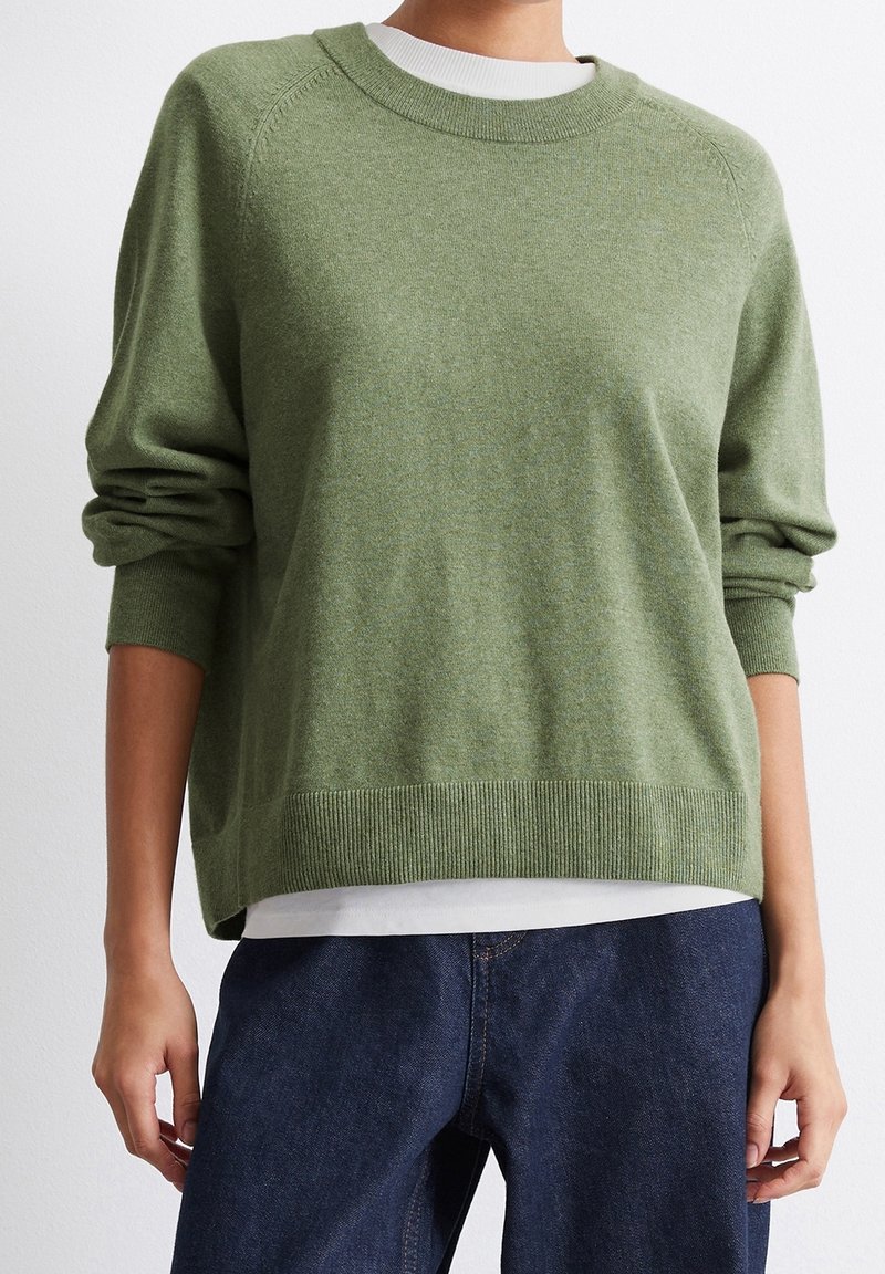 Personne portant un pull à manches longues vert olive par-dessus une chemise blanche, associé à un jean bleu foncé sur fond blanc uni.