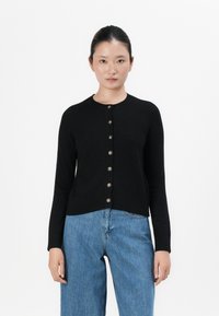 Mulher de pé, de frente, vestindo um cardigan preto com botões e jeans largos de cintura alta azul, em fundo branco.