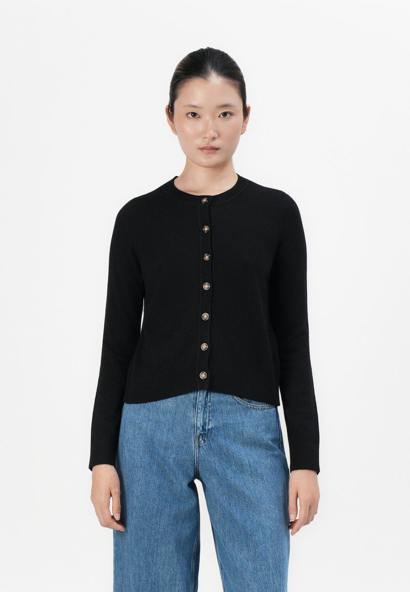 Mulher de pé, de frente, vestindo um cardigan preto com botões e jeans largos de cintura alta azul, em fundo branco.
